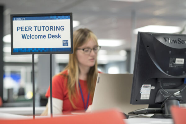 A tutor in a red shirt sits behind the Peer Tutoring Welcome Desk sign
