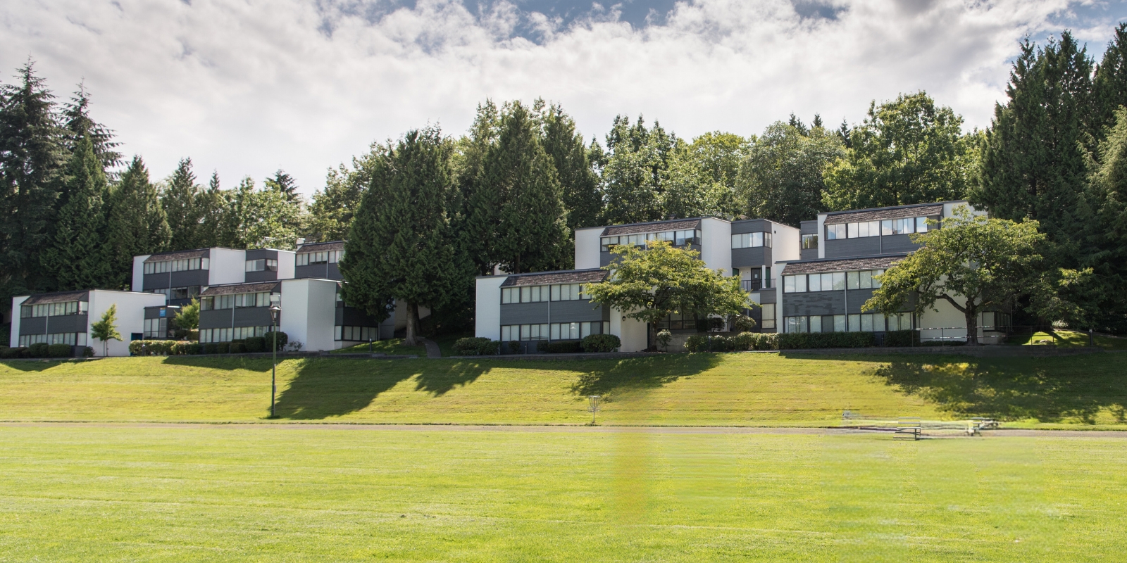 Row of modern, townhouse-style student housing buildings surrounded by lush green trees and a well-maintained grassy field, located on the BCIT Burnaby campus under a partly cloudy sky.